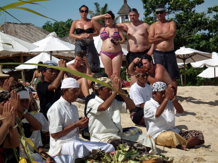 A Bali, des touristes observent une cérémonie religieuse. L'île sera de nouveau ouverte au tourisme international à partir du 11 septembre prochain, malgré la progression de la pandémie de Covid-19 (Image : Wikimedia Commons - Bair175 - https://commons.wikimedia.org/wiki/File:Bali_religi%C3%B6s_ceremoni_p%C3%A5_strand_med_turister.jpg)